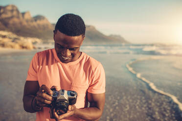 Afrikanischer Mann überprüft Fotos in seiner Digitalkamera. Junger Mann mit digitaler Fotokamera am Strand stehend. - JLPSF00121