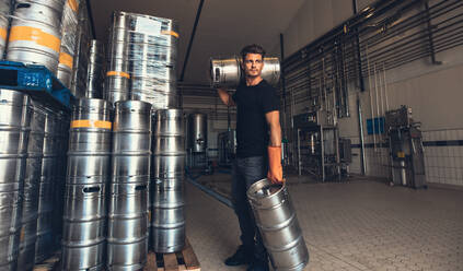 Junger männlicher Brauer, der in einer Brauereifabrik ein Fass trägt. Junger Mann mit Metallbierfässern in einem Lagerhaus. - JLPSF00127