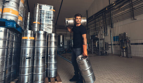 Young male brewer carrying keg at brewery factory. Young man with metal beer barrels at warehouse. - JLPSF00127