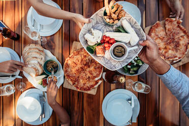 Draufsicht auf Menschen, die an einem für eine Party gedeckten Tisch eine Mahlzeit zu sich nehmen. Freunde feiern eine Einweihungsparty auf einem Holztisch. Männliche Hände reichen einen Teller an einen weiblichen Gast weiter. - JLPSF00132