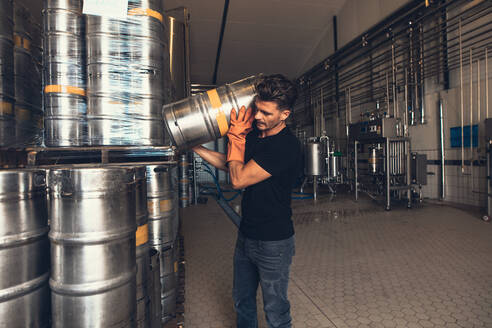 Young male brewer with keg at brewery factory warehouse. Young man working at warehouse in brewery. Arranging metal beer barrel. - JLPSF00133