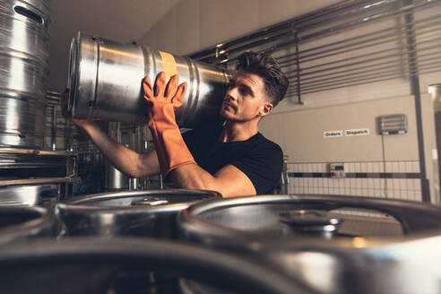 Ein Brauer trägt ein Bierfass aus Metall in einer Brauerei. Ein junger Mann arbeitet in einem Brauereilager. - JLPSF00134