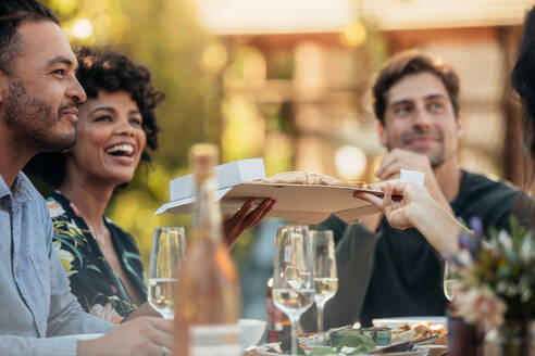 Eine Gruppe junger Freunde sitzt um einen Tisch herum und isst auf einer Party im Freien; sie reichen und servieren ihren Freunden das Essen. - JLPSF00136