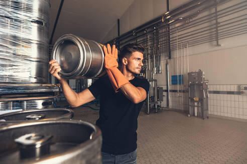 Brewer with large metal container at brewery factory. Young man working at warehouse in brewery. Arranging metal beer barrel at storage area. - JLPSF00138