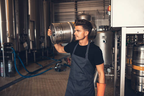 Young male brewer carrying keg at brewery. Manual worker with metal beer barrel craft beer manufacturing plant. - JLPSF00149