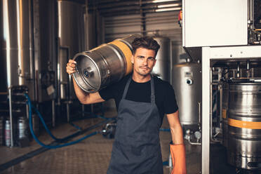 Porträt eines männlichen Brauers, der einen Metallbehälter in einer Brauereifabrik trägt. Junger Mann, der in einem Lagerhaus ein Fass auf der Schulter trägt. - JLPSF00151