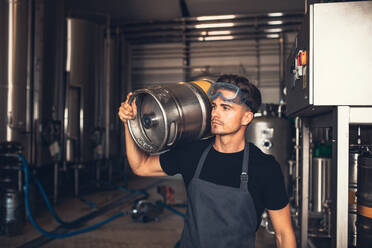 Brauer mit großem Metallbehälter in einer Brauereifabrik. Junger Mann arbeitet im Lager einer Brauerei und hält ein Bierfass aus Metall. - JLPSF00152