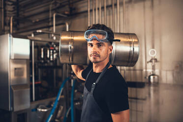 Porträt eines männlichen Brauers mit einem Fass in einer Brauereifabrik. Junger Mann, der in einem Lagerhaus einen Metallbehälter für Bier auf der Schulter trägt. - JLPSF00155