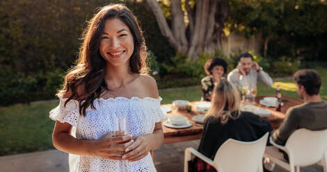 Beautiful asian woman standing outdoors with friends sitting in background having food. Woman with a drinks at outdoor party. - JLPSF00160