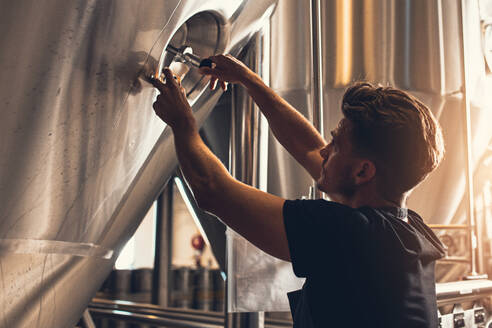 Brewer closing the hatch of brewery tank. Young male employee working in beer manufacturing factory. - JLPSF00166