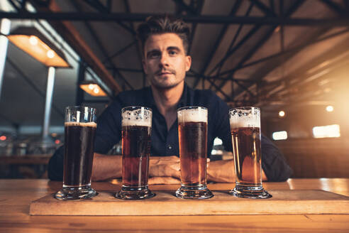 Junger Mann beim Verkosten verschiedener Biersorten auf einem Holztisch in einer Brauerei. Braumeister mit verschiedenen Biersorten am Stehtisch. - JLPSF00177