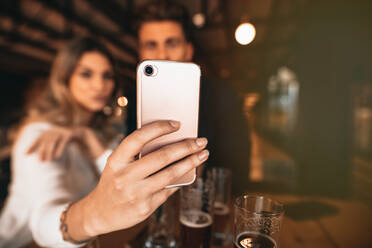 Close up of couple sitting in the bar and taking a selfie with smart phone. Focus on mobile phone in hand of woman. - JLPSF00188