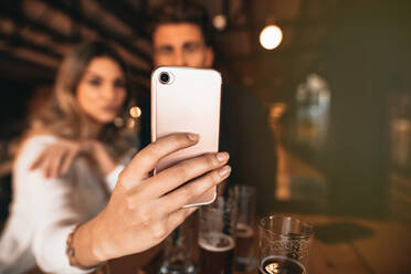 Nahaufnahme eines Paares, das in einer Bar sitzt und ein Selfie mit einem Smartphone macht. Fokus auf das Handy in der Hand der Frau. - JLPSF00188