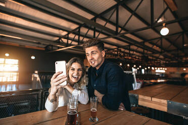 Glückliches Paar, das ein Selfie mit einem Smartphone in einer Bar macht. Ein lächelnder Mann und eine lächelnde Frau sitzen in einer Brauereibar mit Bier auf dem Tisch und machen ein Selbstporträt mit ihrem Mobiltelefon. - JLPSF00189