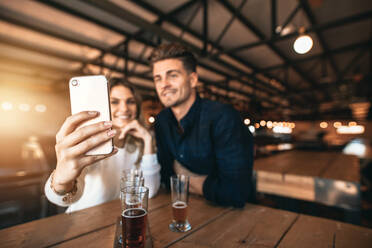 Junges Paar sitzt in der Bar und macht ein Selfie mit dem Handy. Fokus auf das Smartphone in der Hand der Frau. - JLPSF00190