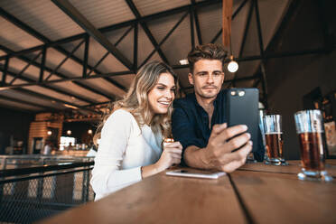 Junges Paar an der Bar, das ein Selfie mit dem Handy macht, Fokus auf das Smartphone in der Hand des Mannes. - JLPSF00192