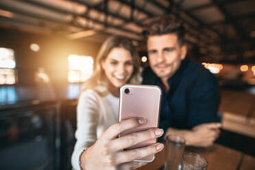 Nahaufnahme eines Paares, das in einer Bar sitzt und ein Selfie mit einem Smartphone macht. Fokus auf das Handy in der Hand der Frau. - JLPSF00193