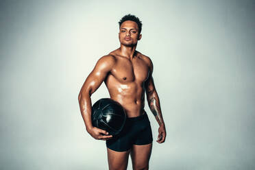 Studioaufnahme eines fitten jungen Mannes mit Medizinball vor grauem Hintergrund. Afrikanisches amerikanisches männliches Modell mit muskulösem Körper, das in die Kamera schaut. - JLPSF00198