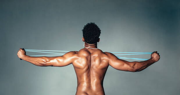 Rear view of muscular young man stretching arms with rubber band. Male bodybuilder doing exercises with elastic band on grey background. - JLPSF00199