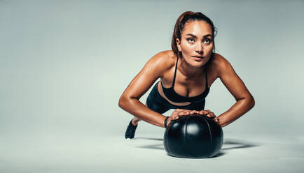 Horizontale Aufnahme einer jungen, fitten Frau, die einen Liegestütz mit einem Medizinball macht, auf einem grauen Hintergrund. - JLPSF00207