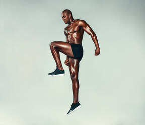 Full length portrait of a fitness man jumping and stretching over grey background. African fitness model with muscular build exercising. - JLPSF00221