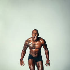 Muskulöser junger Mann schreit vor grauem Hintergrund. Hemdfreies männliches Model mit muskulösem Körperbau, das schreit. - JLPSF00226