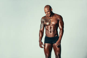 Porträt eines muskulösen jungen Mannes, der vor einem grauen Hintergrund lächelt. Ein afroamerikanisches Fitnessmodell blickt nach unten und lächelt. - JLPSF00228