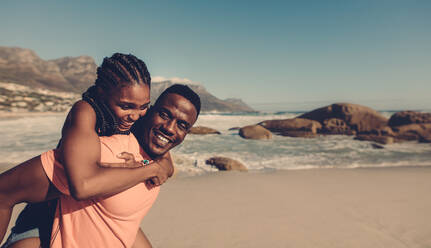 Junger afrikanischer Mann, der seine Freundin am Strand auf dem Rücken trägt. Verliebtes Paar, das sich am Meer huckepack vergnügt. - JLPSF00250