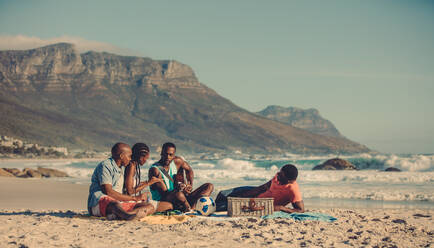 Ein afrikanischer Mann spielt Gitarre für seine Freunde am Meer. Eine Gruppe von Menschen entspannt sich am Sandstrand. - JLPSF00280