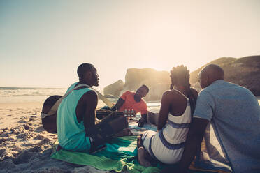 Afrikanischer junger Mann, der am Strand singt und Gitarre spielt, Gruppe von vier Personen, die sich beim Picknick am Strand amüsieren. - JLPSF00281