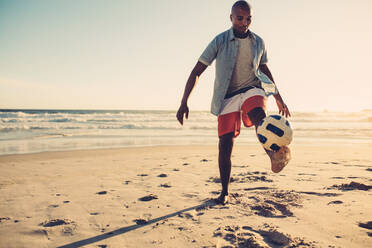 Junger Mann spielt Fußball am Meer. Afrikanischer Mann kickt Fußball am Strand. - JLPSF00282