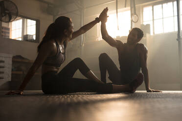 Gesunder Mann und Frau sitzen auf dem Boden und geben sich gegenseitig High Five im Fitnessstudio. Fitness Menschen nach erfolgreichem Training im Fitnessstudio. - JLPSF00300