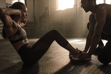 Sportlerin macht Sit-ups, während ein Fitnesstrainer ihre Füße hält. Frau macht Bauchmuskelübungen mit Hilfe eines Mannes im Fitnessstudio. - JLPSF00302