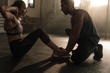 Männlicher Trainer, der einer Frau bei Sit-ups im Fitnessstudio hilft. Eine Frau trainiert ihre Bauchmuskeln mit Hilfe eines männlichen Personal Trainers. - JLPSF00304