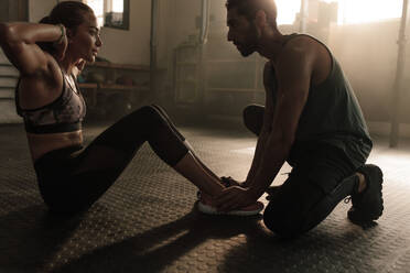 Male coach assisting woman in doing sit-ups at gym. Woman working out on her abs with help from male personal trainer. - JLPSF00304