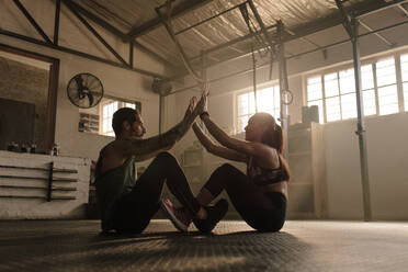 Ein Paar, das zusammen im Fitnessstudio trainiert. Ein fitter Mann und eine fitte Frau sitzen auf dem Boden und geben sich beim Training ein High Five. - JLPSF00308