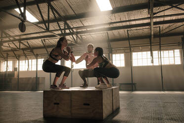 Fitte junge Leute, die gemeinsam in einer Turnhalle Boxsprünge machen. Trainingsgruppe, die im Fitnessstudio auf Holzkisten springt. - JLPSF00322