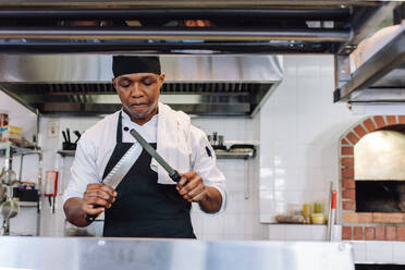 Afrikanischer Koch schärft sein Messer in einer Restaurantküche. Koch bereitet sich auf das Kochen in einer Großküche vor. - JLPSF00349