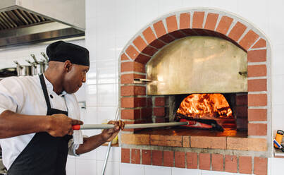 Koch backt Pizza im Holzofen in einer Großküche. Koch backt Pizza in einem traditionellen Steinofen in einem Restaurant. - JLPSF00357