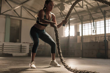 Junge Frau beim Krafttraining mit Kampfseilen im Fitnessstudio. Die Athletin bewegt die Seile in einer Wellenbewegung als Teil des Fettverbrennungstrainings. - JLPSF00358