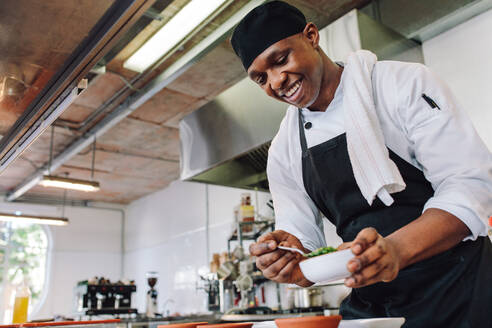 Gourmetkoch in Uniform, der in einer Großküche kocht. Glücklicher männlicher Koch mit Schürze, der an der Küchentheke steht und Essen zubereitet. - JLPSF00363