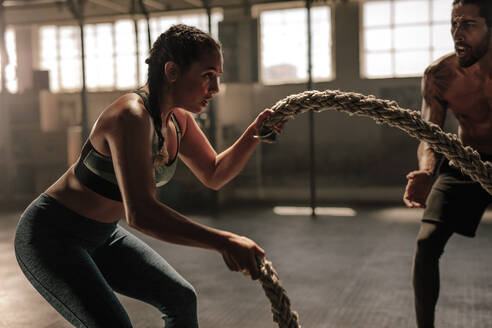 Starke Frau trainiert mit Battle Rope im Fitnessstudio mit männlichem Trainer. Athlet macht Battle Rope Workout im Fitnessstudio mit Instruktor. - JLPSF00367