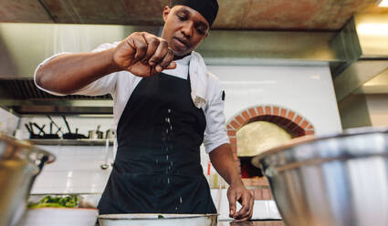 Afrikanischer männlicher Koch, der in einer Restaurantküche Essen zubereitet und Gewürze hinzufügt. - JLPSF00369