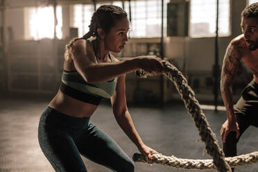 Frau trainiert mit Battle Ropes im Fitnessstudio mit Trainer. Athlet macht Battle Rope Workout im Fitnessstudio mit Trainer. - JLPSF00375