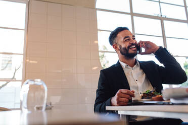 Lächelnder junger Geschäftsmann sitzt am Restauranttisch mit Essen und spricht mit seinem Handy. Kaukasischer Geschäftsmann im Restaurant beim Telefonieren. - JLPSF00393