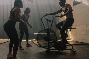 Two women motivating man exercising on air bike in gym. Man doing intense workout on gym bike with females. - JLPSF00400