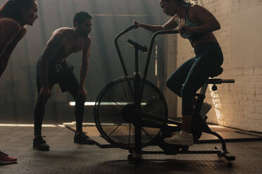 Fitness woman on bicycle doing spinning at gym with trainers. Fit young female working out on gym bike with young people. - JLPSF00407