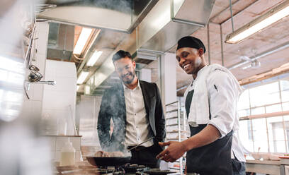 Lächelnder Koch, der Essen auf dem Herd zubereitet, während der Restaurantleiter daneben steht. Restaurantleiter mit Koch in der Großküche. - JLPSF00411