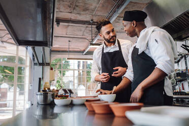 Zwei männliche Köche bereiten Essen in einer Restaurantküche zu. Köche unterhalten sich beim Kochen von Essen in einer Großküche. - JLPSF00419