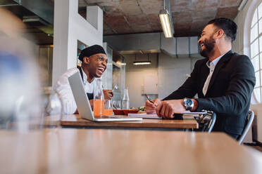 Lächelnder Restaurantleiter, der am Tisch sitzt und sich mit dem Chefkoch unterhält. Restaurantbesitzer, der ein freundliches Gespräch mit einem Mitarbeiter führt. - JLPSF00458